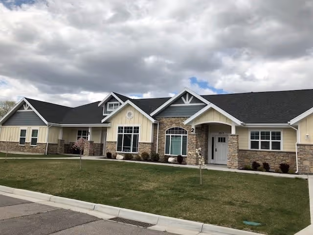 Exterior view of a single-story building with a combination of stone and light yellow siding, multiple windows, and a dark shingled roof under a cloudy sky. The building is surrounded by a well-maintained lawn with a few small trees and a sidewalk in front.