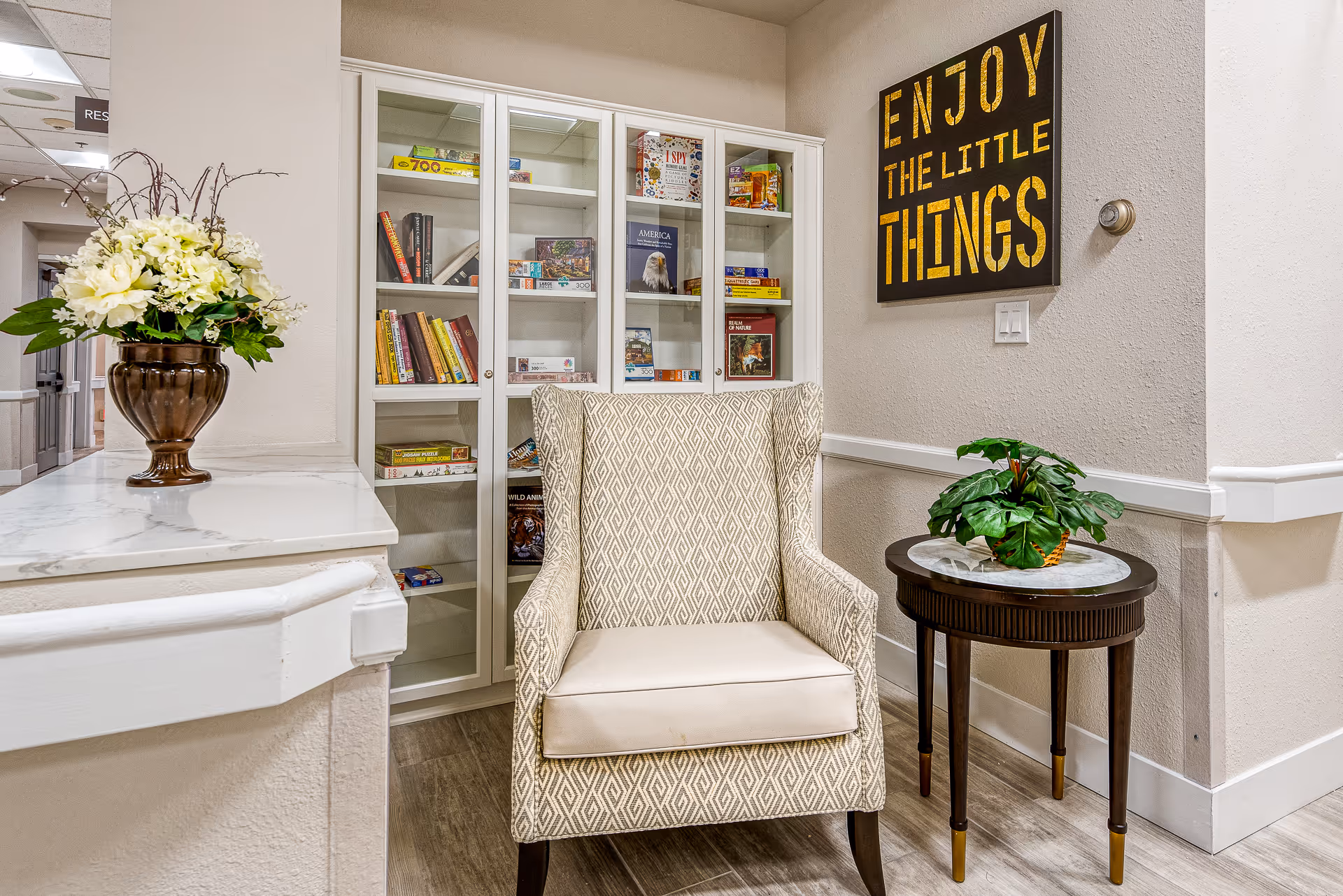 Upholstered armchair beside a small round side table and a glass-front bookcase under a wall sign that reads 'Enjoy the little things'.