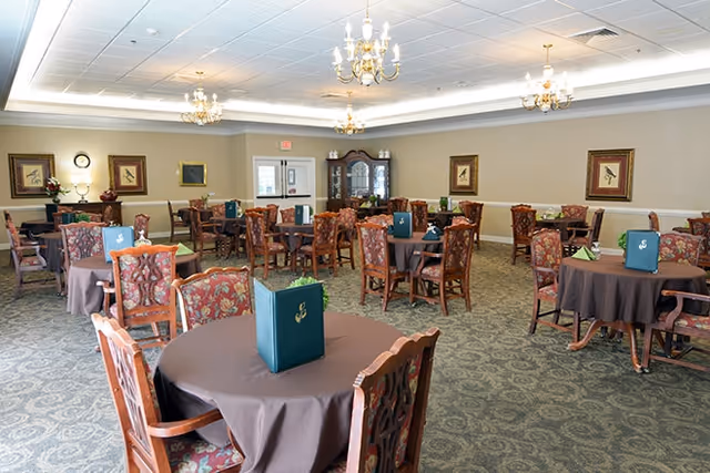 A spacious dining room with multiple round tables covered with brown tablecloths, each surrounded by wooden chairs with floral upholstery. Menus are placed on each table. The room is well-lit with chandeliers hanging from the ceiling and decorated with framed artwork on the walls. A wooden cabinet and a piano are visible in the background.
