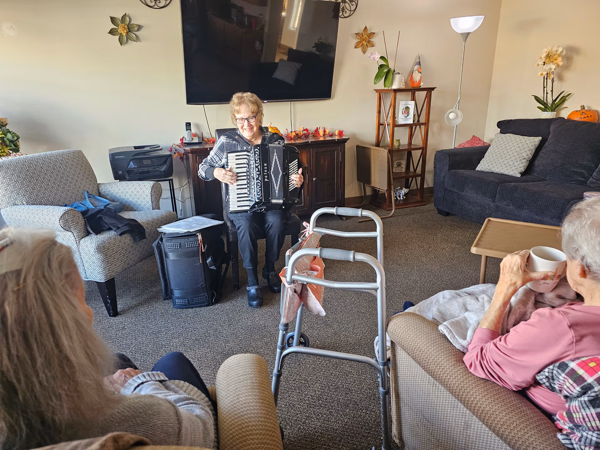 An elderly woman plays an accordion in a communal living room while other residents sit and watch.