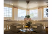 A bright and airy sitting area with two beige armchairs and a glass coffee table adorned with a small bouquet of yellow flowers and magazines. Large windows let in natural light, and a decorative plant sits on a ledge behind the chairs. The space features soft neutral tones and elegant architectural details.