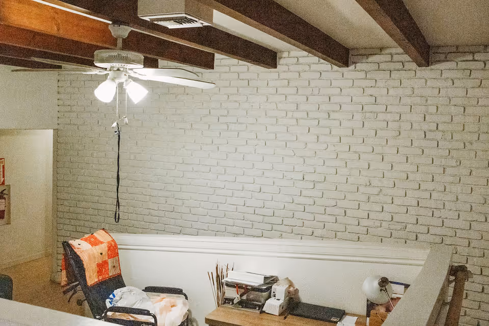Loft-style sitting area with a ceiling fan, exposed wooden beams, a painted brick wall, and a chair with a quilt beside a small table and lamp.