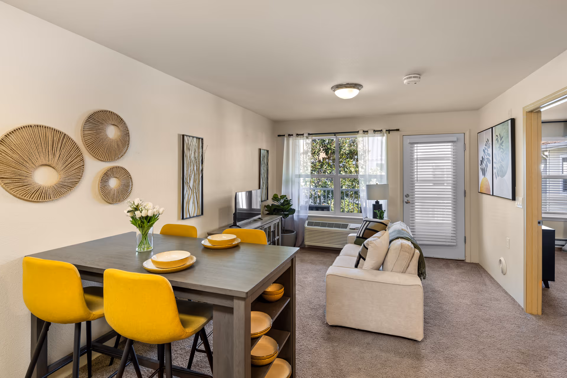 A bright and cozy living room and dining area in a senior living facility. The dining area features a gray table with four yellow chairs and place settings with yellow plates. On the wall above the table are three decorative circular wall hangings. The living room has a beige sofa with cushions, a TV on a stand, a potted plant, and a window with sheer white curtains letting in natural light. A door with blinds is next to the window, and two framed botanical prints hang on the wall near an open doorway leading to another room.