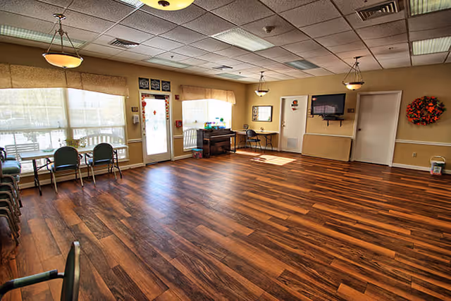A spacious room with wood flooring and beige walls, featuring large windows with blinds and valances, several chairs stacked and arranged along the walls, a wall-mounted television, a small desk with a chair, and a door leading outside. The ceiling has recessed lighting and hanging light fixtures.