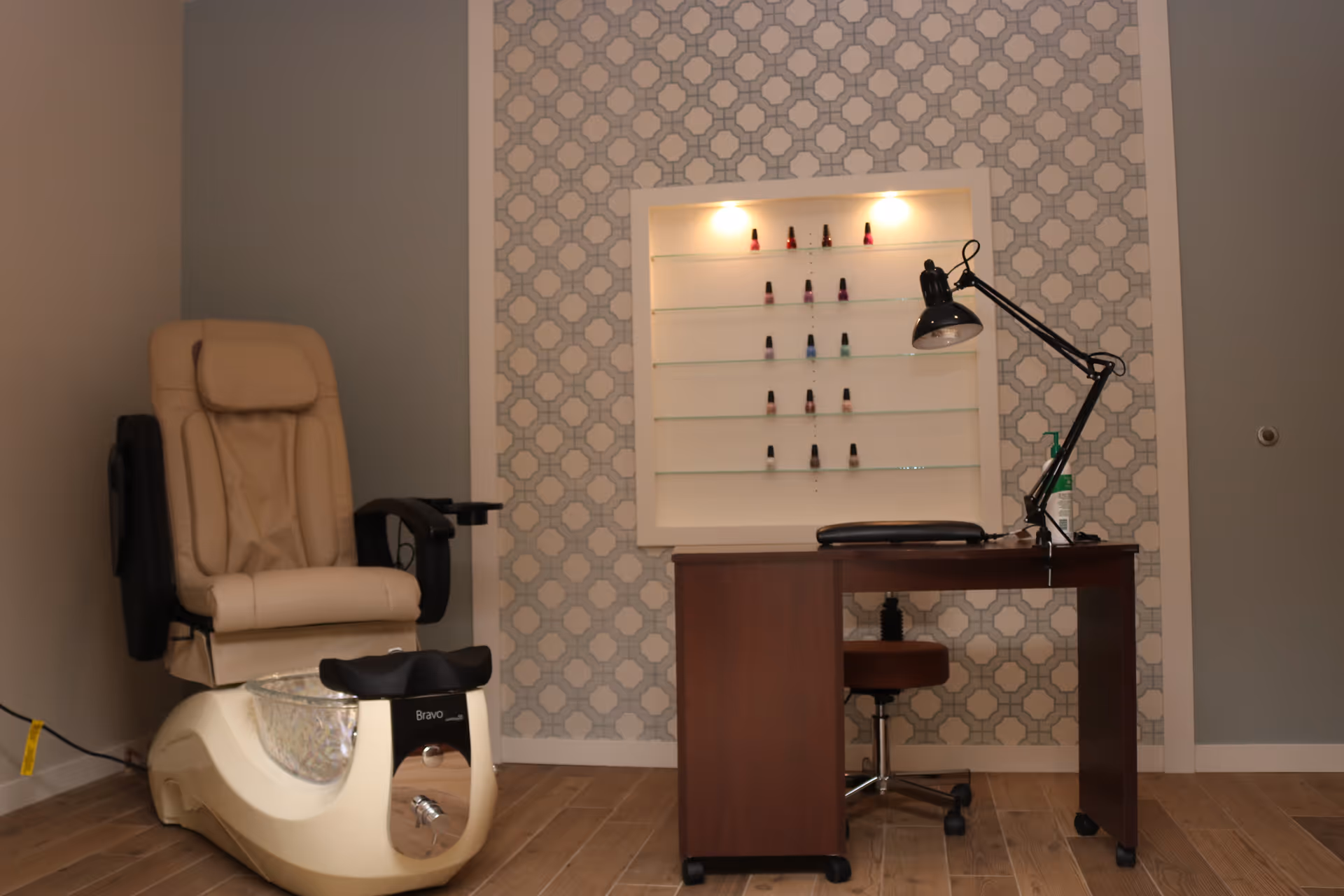 A nail salon station with a beige pedicure chair and foot bath on the left, and a wooden manicure table with a black adjustable lamp on the right. Behind the table is a wall with patterned wallpaper and a display case with various nail polish bottles.