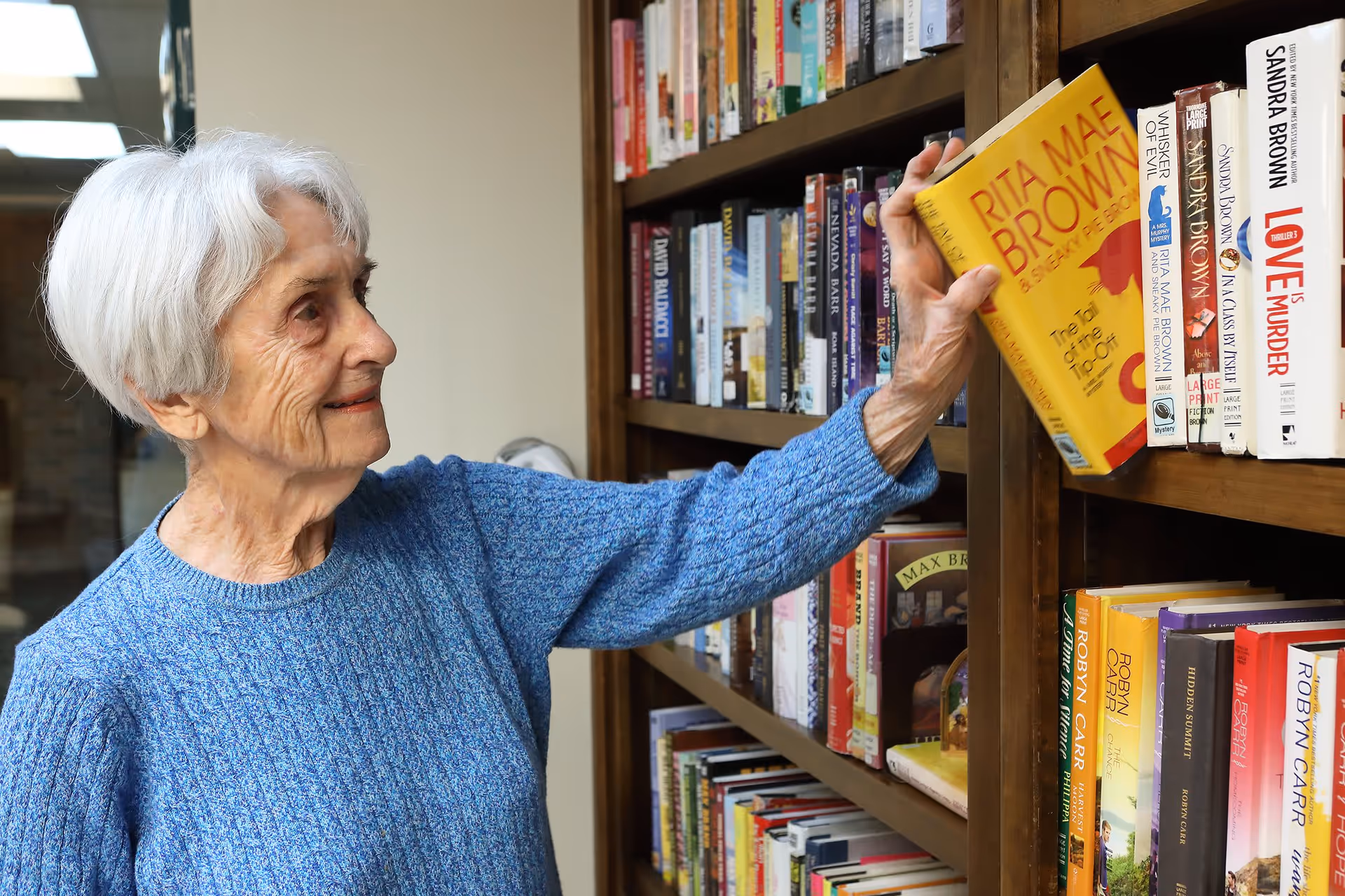 An elderly woman with short white hair wearing a blue sweater is reaching for a yellow book titled 'Rita Mae Brown' from a wooden bookshelf filled with various books.