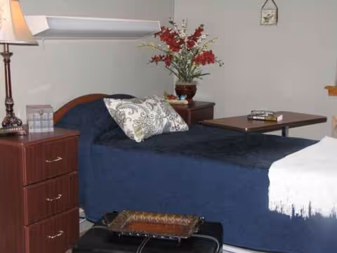 A neatly made bedroom with a blue bedspread, wooden dresser, and red floral arrangement.