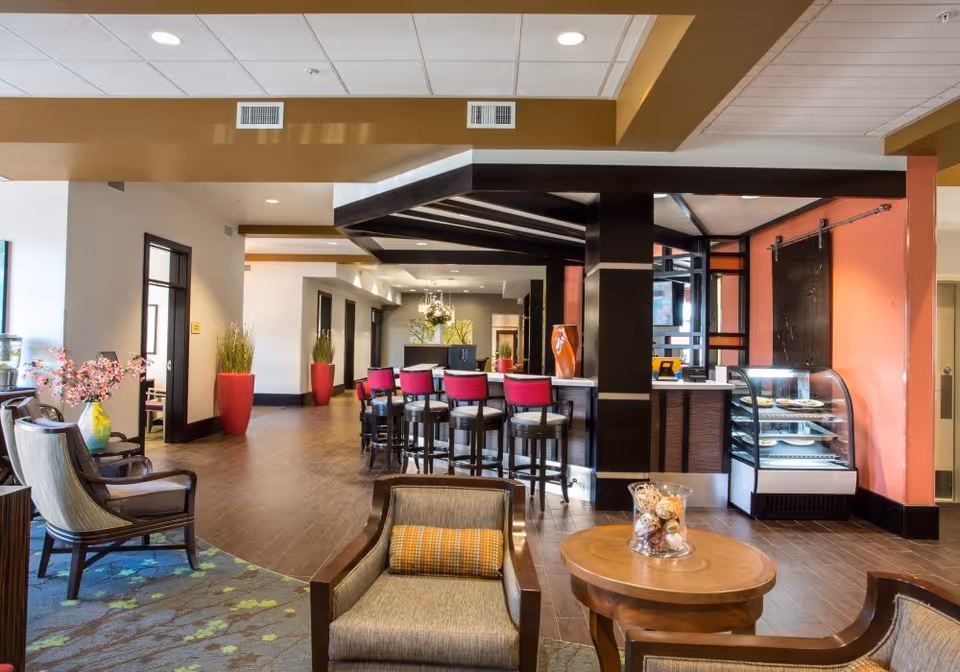 A modern and spacious common area in a senior living facility featuring comfortable armchairs with cushions, a round wooden table with decorative items, a bar counter with high stools that have red cushions, and a display case with pastries. The room has wood flooring, a patterned carpet, large potted plants, and warm lighting with a mix of white and coral-colored walls.