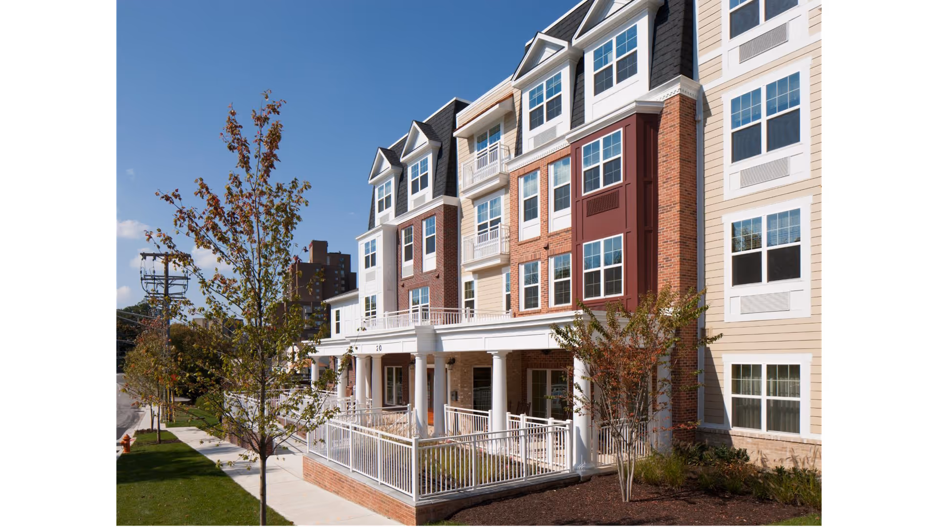 Exterior front of a multi-story senior living building with brick and siding facade, white columns, porches, trees and a sidewalk.