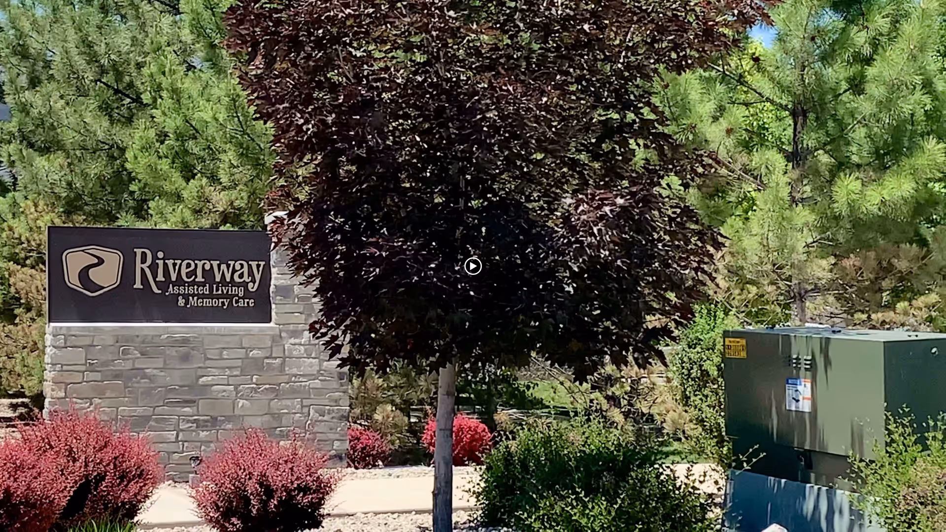 Outdoor view of the entrance sign for Riverway Assisted Living and Memory Care surrounded by trees and bushes.
