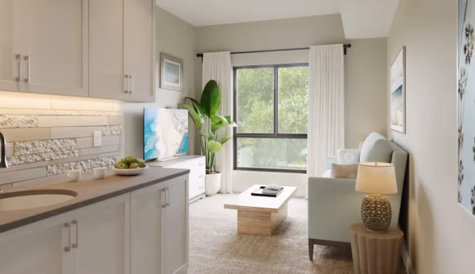 A bright and modern living room area with a large window showing green trees outside. The room features a light blue sofa with pillows, a wooden coffee table with a tray, a side table with a lamp, a flat-screen TV on a white TV stand, and a tall green plant next to the window. The foreground shows a kitchen counter with a bowl of green apples and two small cups, with light-colored cabinets and a textured backsplash.
