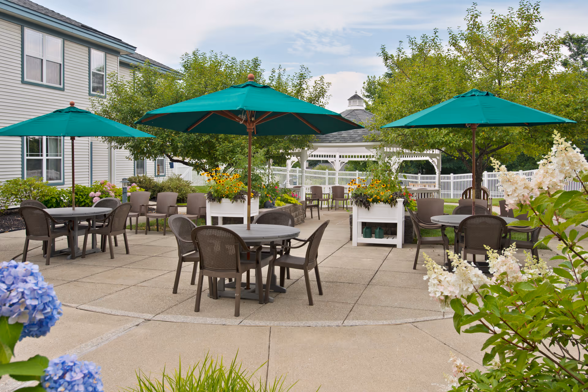 Outdoor patio area at a senior living facility with multiple round tables and chairs under green umbrellas. The patio is surrounded by greenery, flowers, and trees, with a white gazebo and a white picket fence in the background. The building exterior is visible on the left side.
