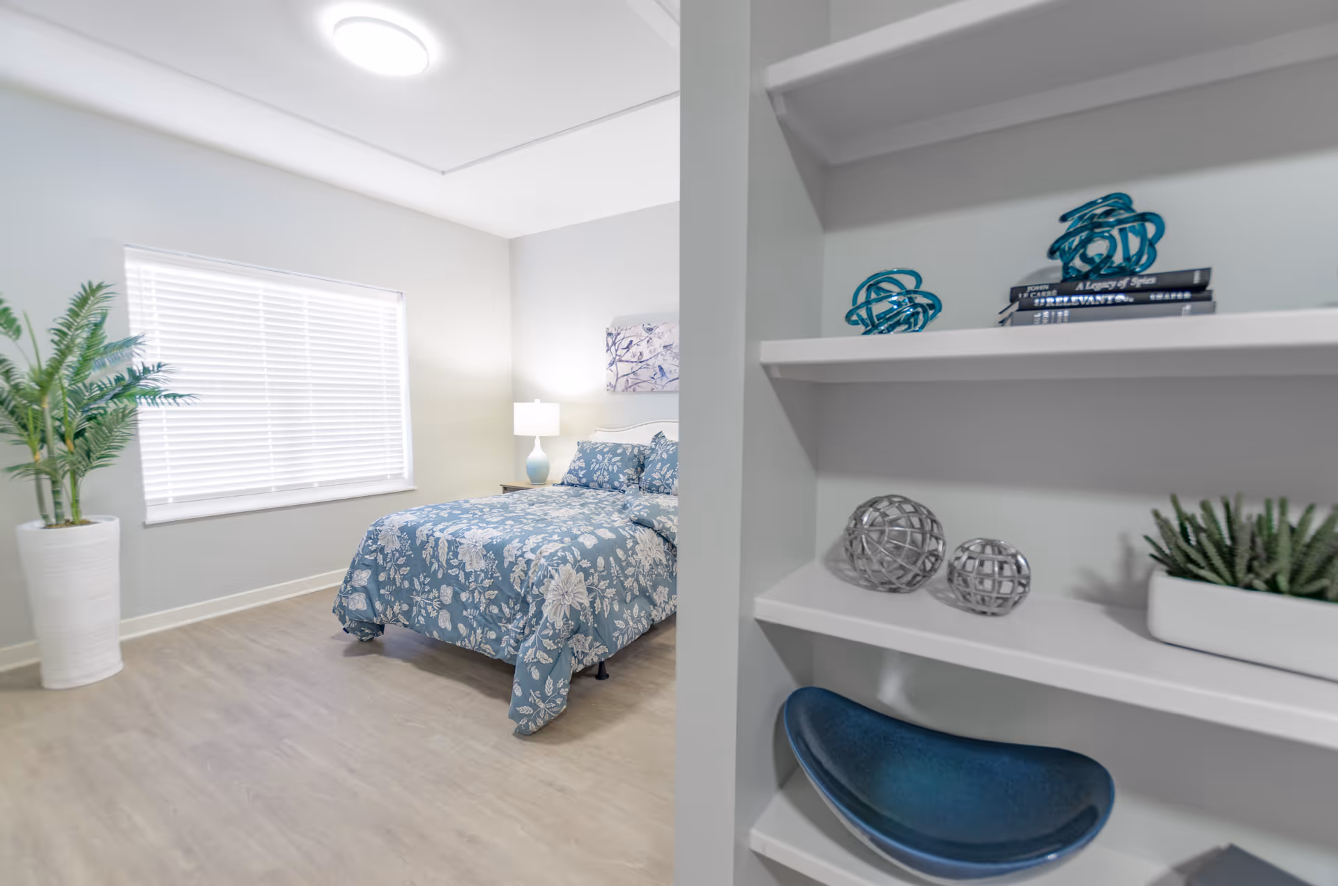 A bright bedroom with a bed covered in blue floral bedding, a bedside table with a lamp, a large window with white blinds, a tall potted plant, and a white shelving unit with decorative items including blue and silver spheres and a small plant.