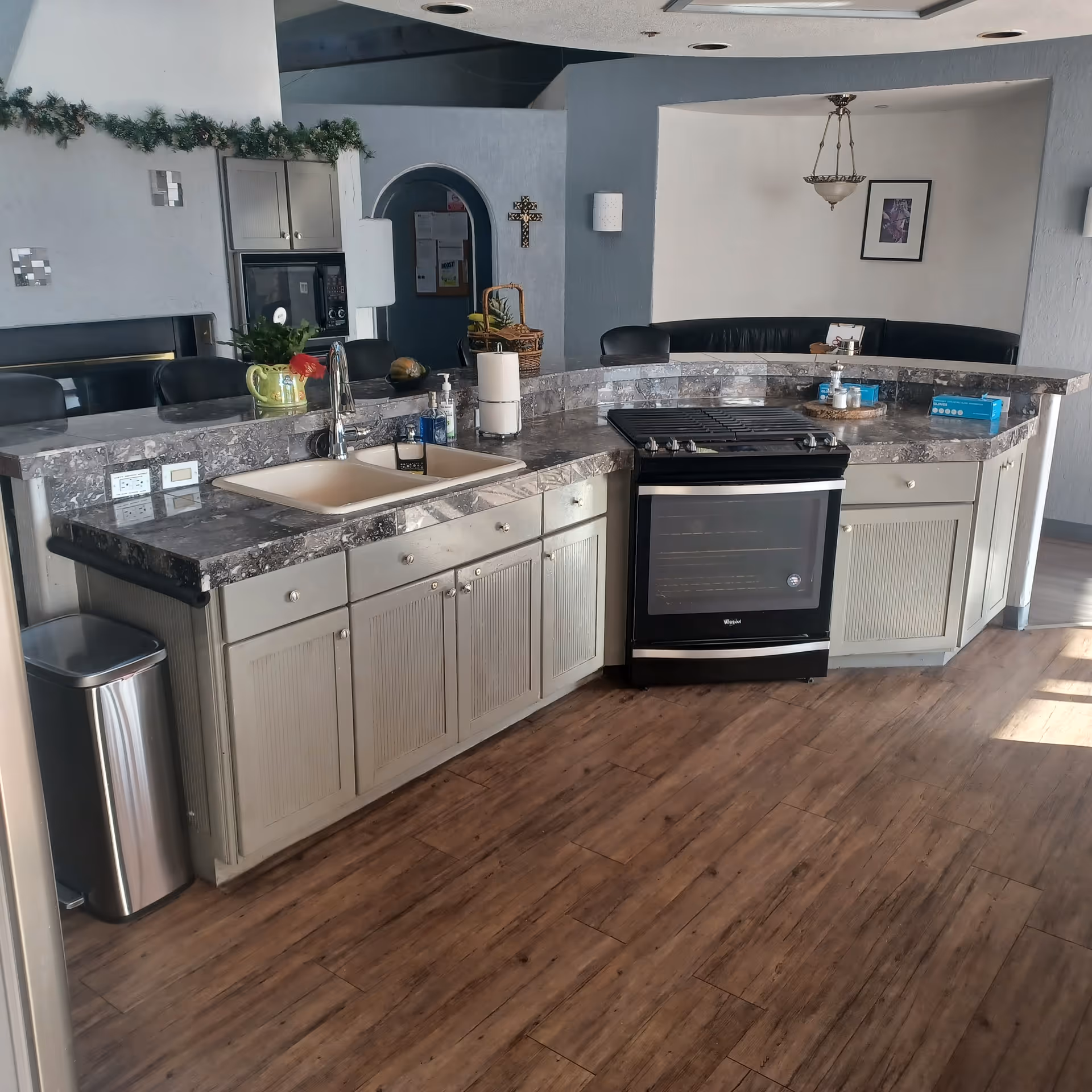 A kitchen area with a marble countertop, a sink, a stove, and cabinets. There is a trash can to the left, a basket with fruit on the counter, and a few small items like paper towels and soap near the sink. In the background, there is a dining nook with a curved black bench and a hanging light fixture, along with a framed picture on the wall.