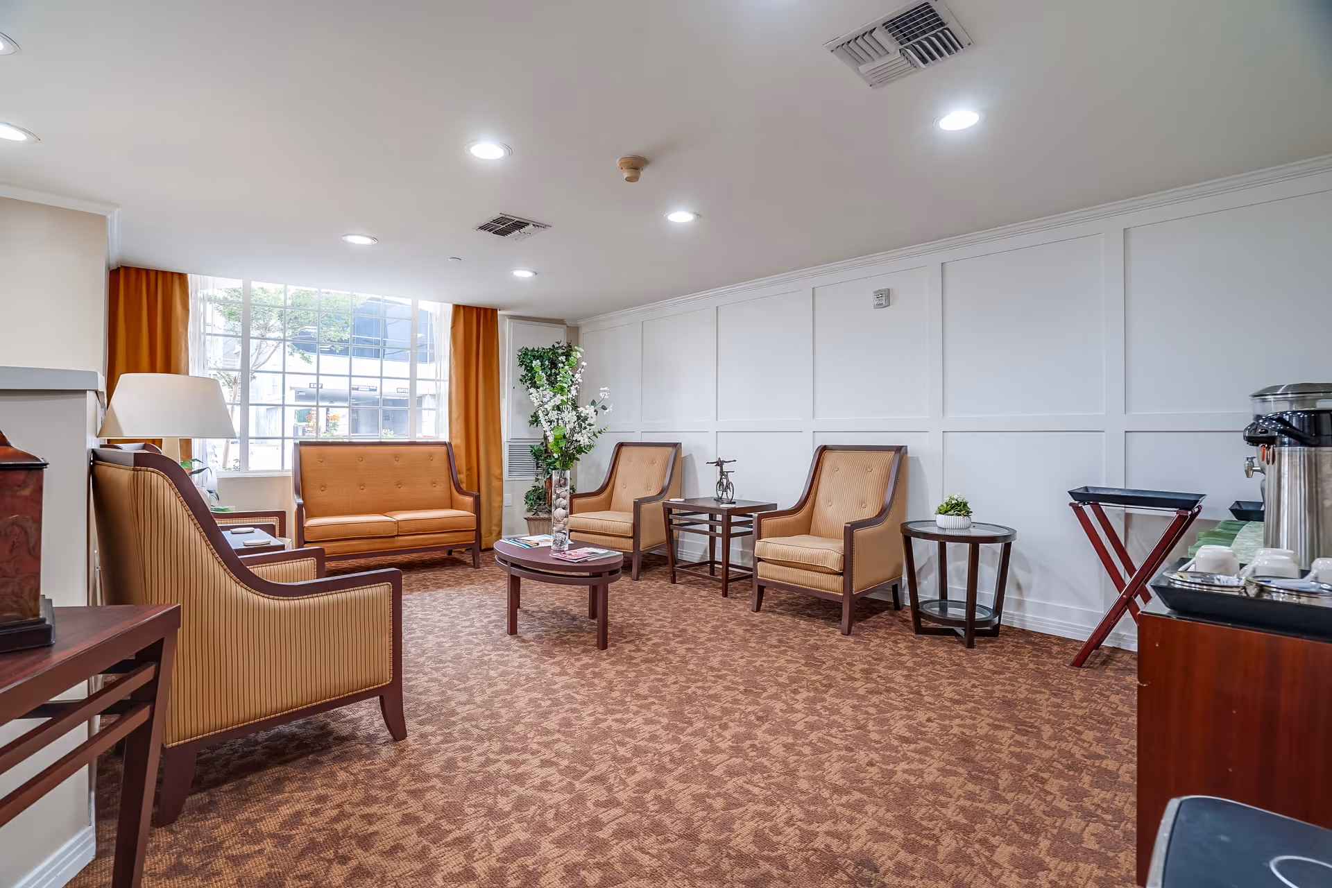 Well-lit senior living lounge with upholstered sofas and chairs arranged around coffee tables near a large window.