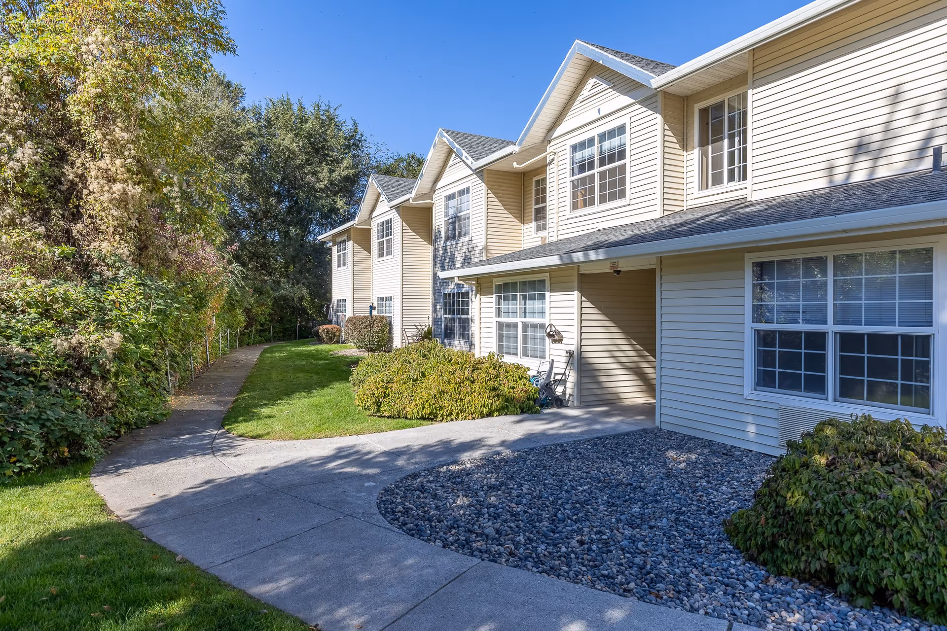 Exterior view of a beige two-story assisted living facility building with multiple windows, surrounded by green bushes and trees under a clear blue sky. A curved concrete pathway leads alongside the building through a landscaped area with grass and rocks.
