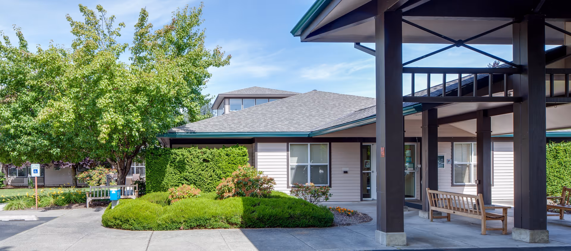 Exterior view of Linden Grove Health Care Center showing a covered entrance with wooden benches, a landscaped area with bushes and trees, and a clear blue sky.