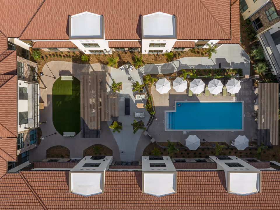 Aerial view of a courtyard showing a rectangular swimming pool, lounge chairs with umbrellas, seating areas and surrounding buildings.