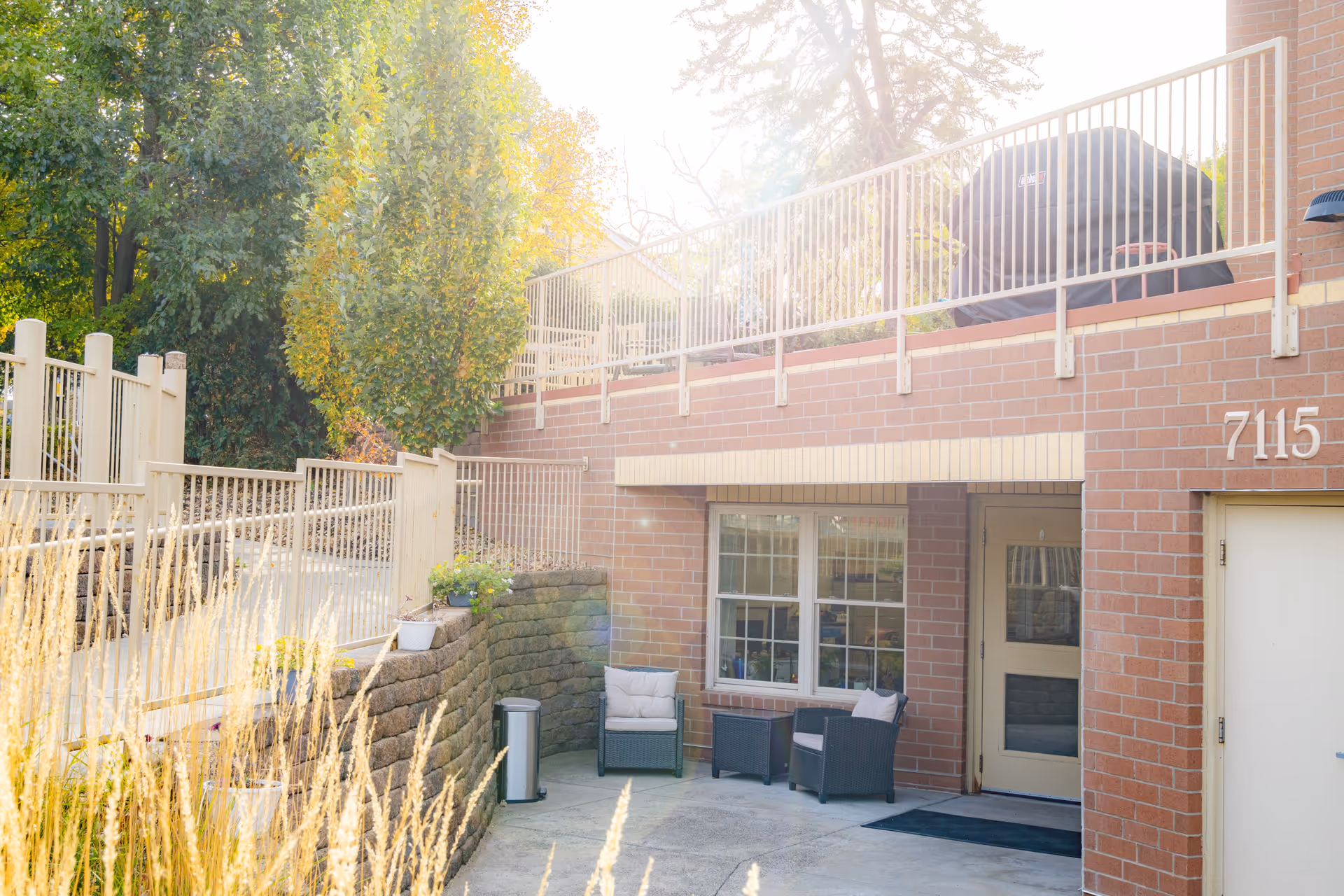 Outdoor patio area at Cedar Ridge Place with two cushioned chairs and a small table set against a brick wall. There is a door and window on the building, a grill on the upper deck, and trees and plants surrounding the space.