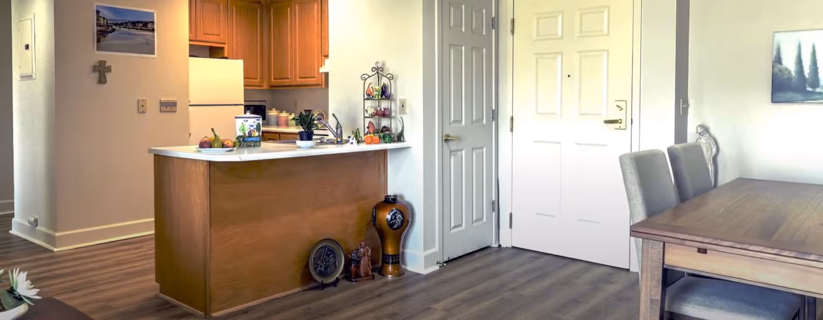 Interior view of a retirement living community showing a kitchen area with wooden cabinets, a white refrigerator, and a countertop with various items including fruit and plants. Adjacent to the kitchen is a dining area with a wooden table and upholstered chairs. The floor is wood laminate, and there are two closed doors visible on the wall opposite the kitchen. The walls are light-colored, decorated with a cross and a framed picture.