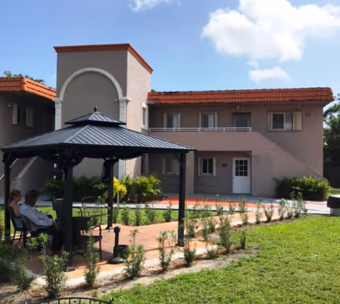 Outdoor courtyard area of Green Life Assisted Living Facility with a black metal gazebo, several chairs, and two people sitting under the gazebo. The building in the background is two stories with a beige exterior and red-tiled roof. The courtyard has a paved walkway and small plants along the path, with a grassy lawn surrounding the area.