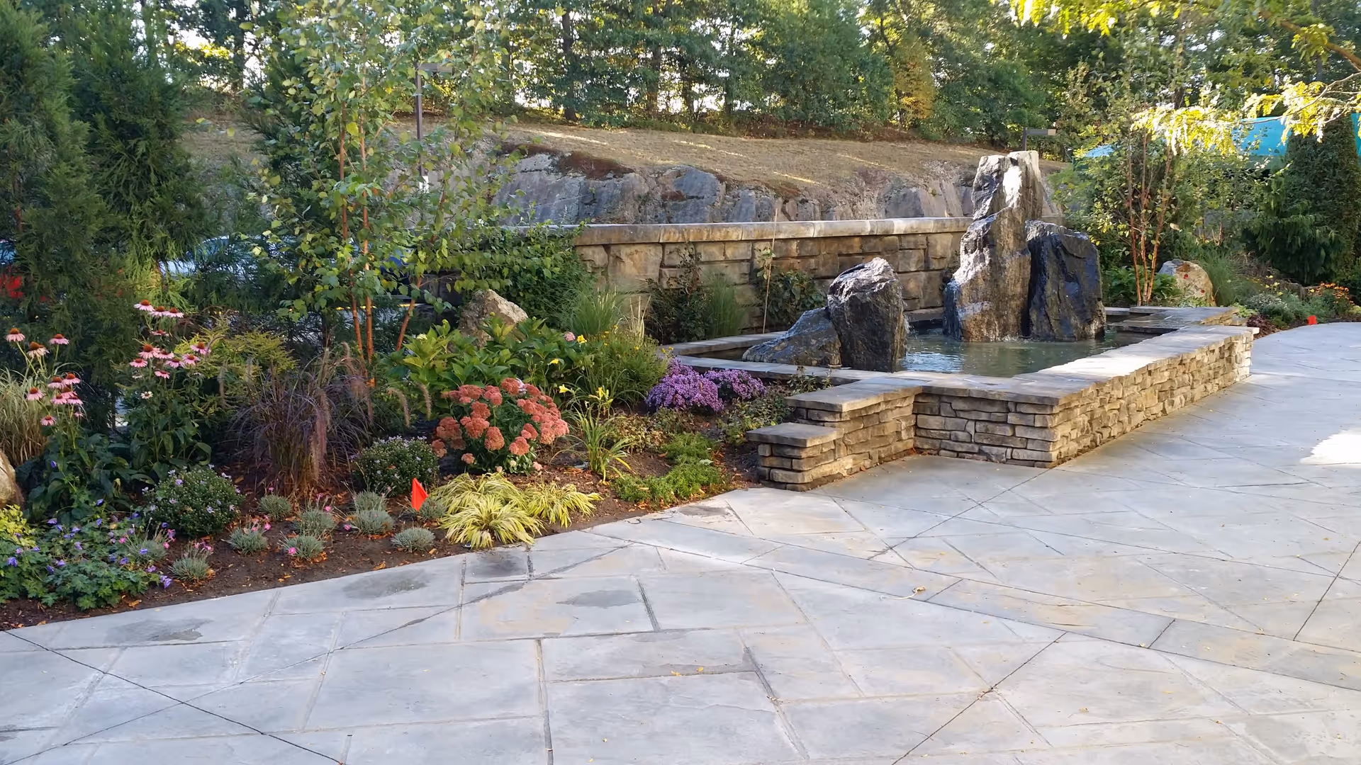 Outdoor garden area with a stone water feature consisting of large rocks in a rectangular stone basin filled with water. Surrounding the water feature are various green plants and colorful flowers, with a stone wall and trees in the background. The foreground shows a paved stone walkway.