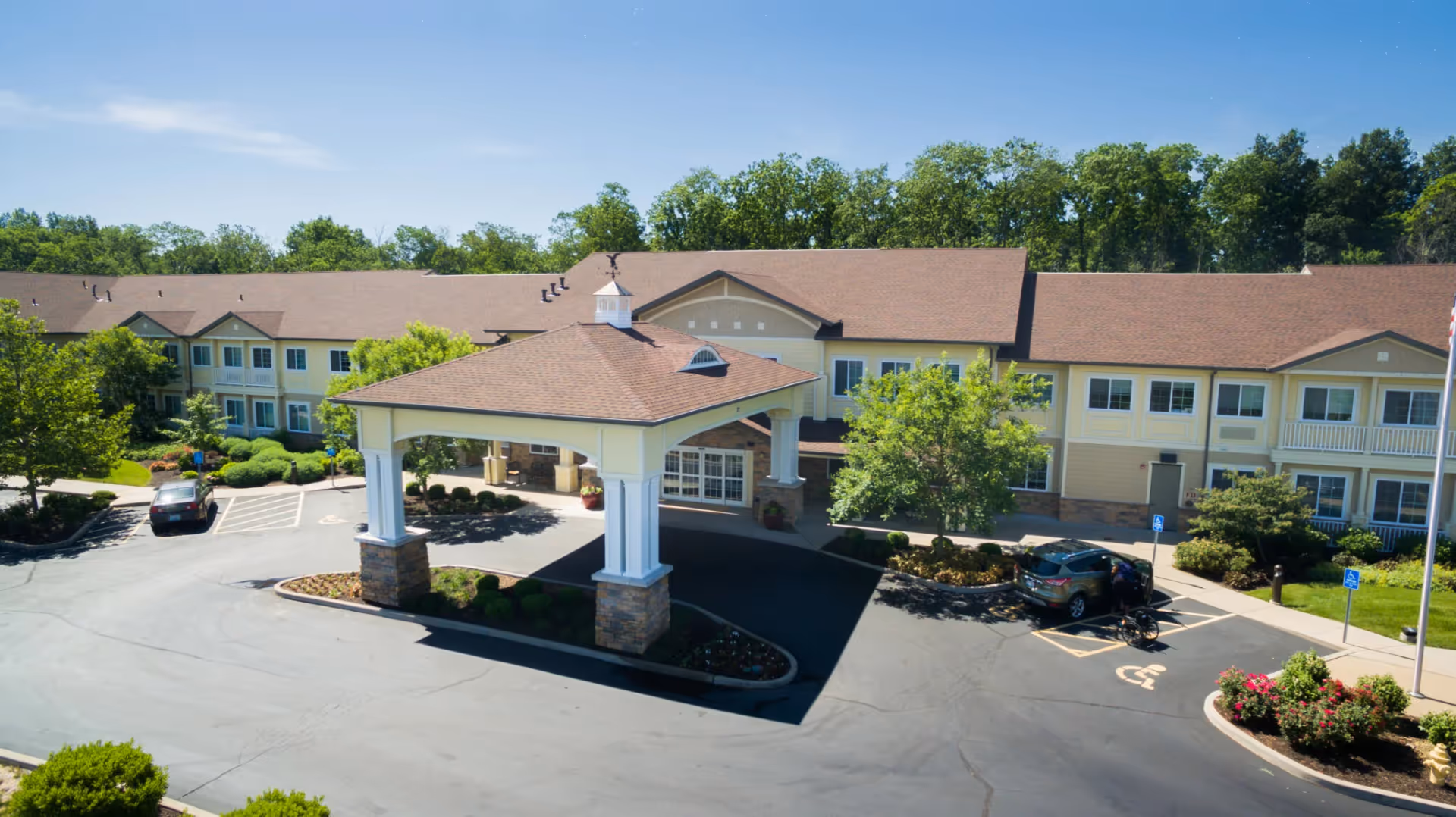 Front view of a two-story assisted living building with a covered porte-cochère, driveway, parked cars, and landscaped grounds.