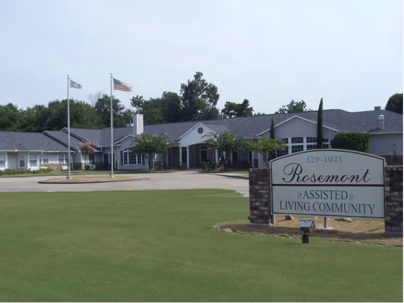 Front exterior view of a single-story assisted living community building with a large green lawn in front, two flagpoles with flags, and a sign that reads 'Rosemont Assisted Living Community'.