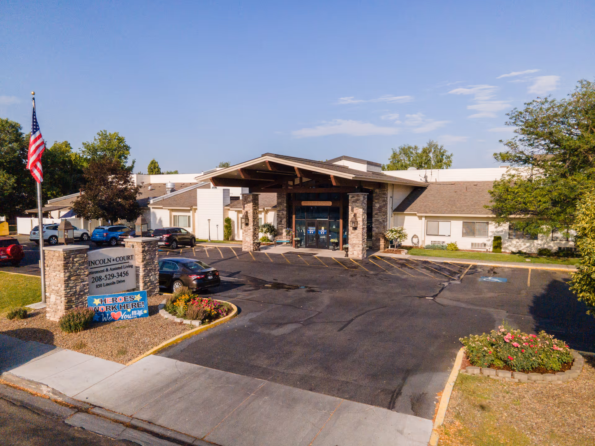 Front exterior view of Lincoln Court Assisted & Senior Living facility with a parking lot, an American flag, and a sign that reads 'Heroes Work Here! We Love You!!!' under the main sign displaying the facility's name and contact information.