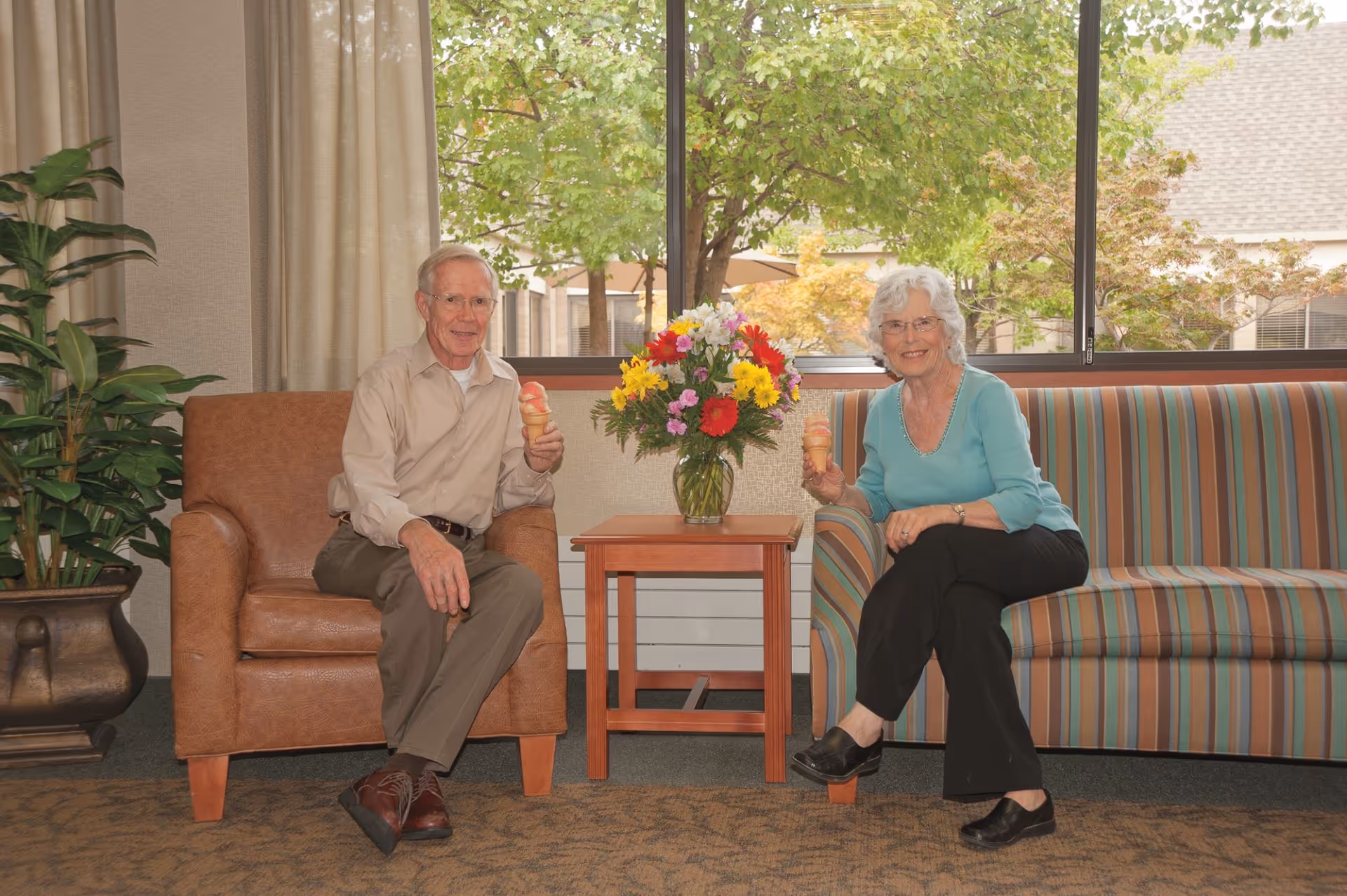 An elderly man and woman sitting in a living room area, each holding an ice cream cone. The man is seated on a brown armchair, and the woman is seated on a striped couch. Between them is a wooden side table with a vase of colorful flowers. Behind them is a large window showing green trees outside.