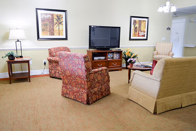 A cozy living room area with a beige couch, two patterned armchairs, a wooden TV stand with a flat-screen television, a side table with a lamp and plant, and framed artwork on the walls.