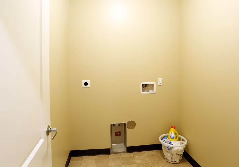 Small beige laundry/utility room with washer/dryer hookups and a laundry basket with detergent.
