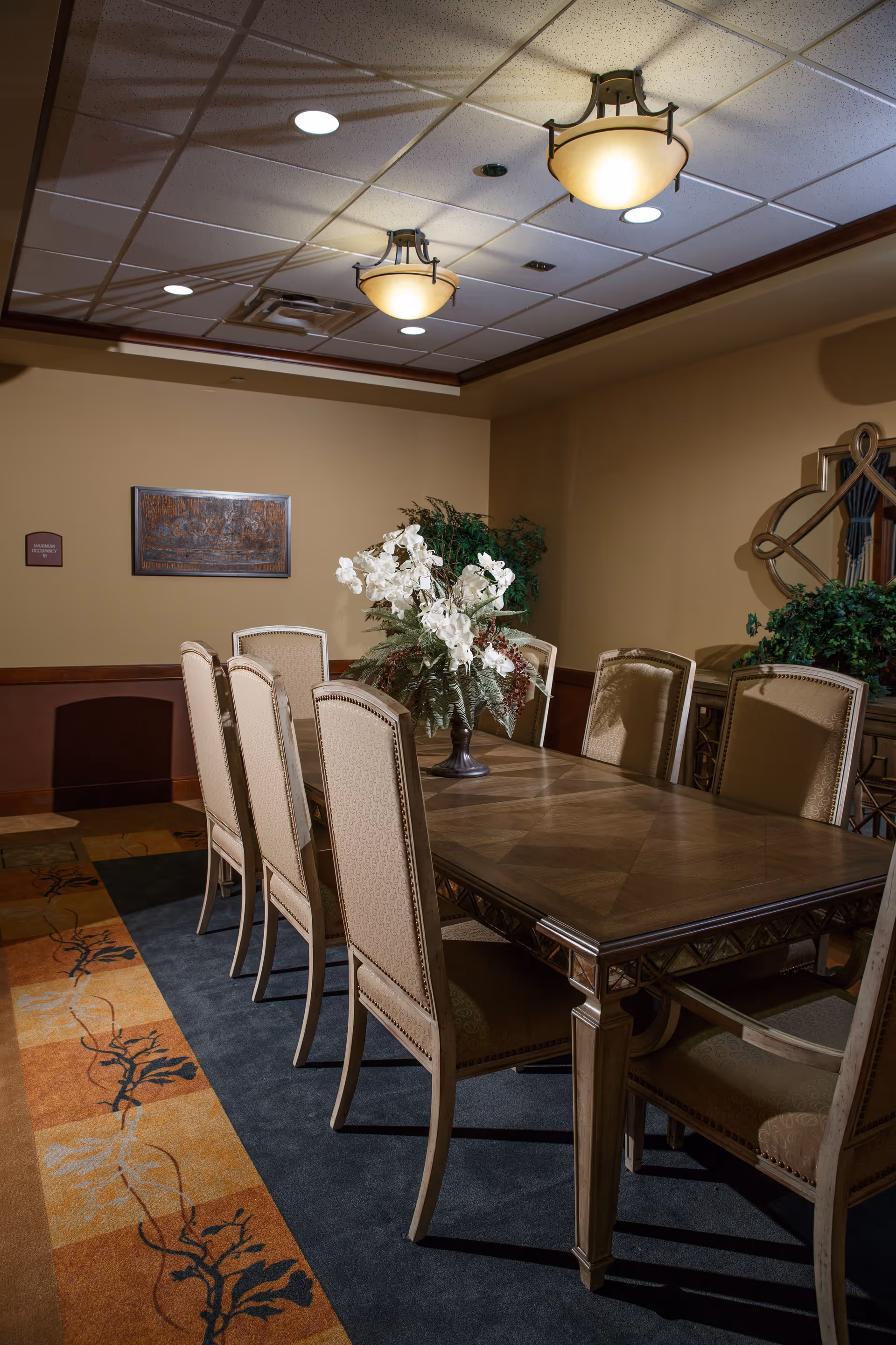 A formal dining room with a long wooden table surrounded by eight upholstered chairs. A floral arrangement with white flowers and greenery is placed in the center of the table. The room has beige walls, a decorative mirror on one wall, a framed artwork, and a patterned carpet with orange and dark blue colors. Ceiling lights illuminate the space.