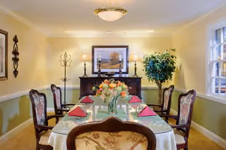 A formal dining room with a rectangular table covered with a white tablecloth and set with red napkins. The table has a floral centerpiece. There are six upholstered chairs around the table. The room has light yellow walls with white trim, a window on the right side, a wooden sideboard with two lamps and a framed painting above it, a coat rack, and a potted plant in the corner.