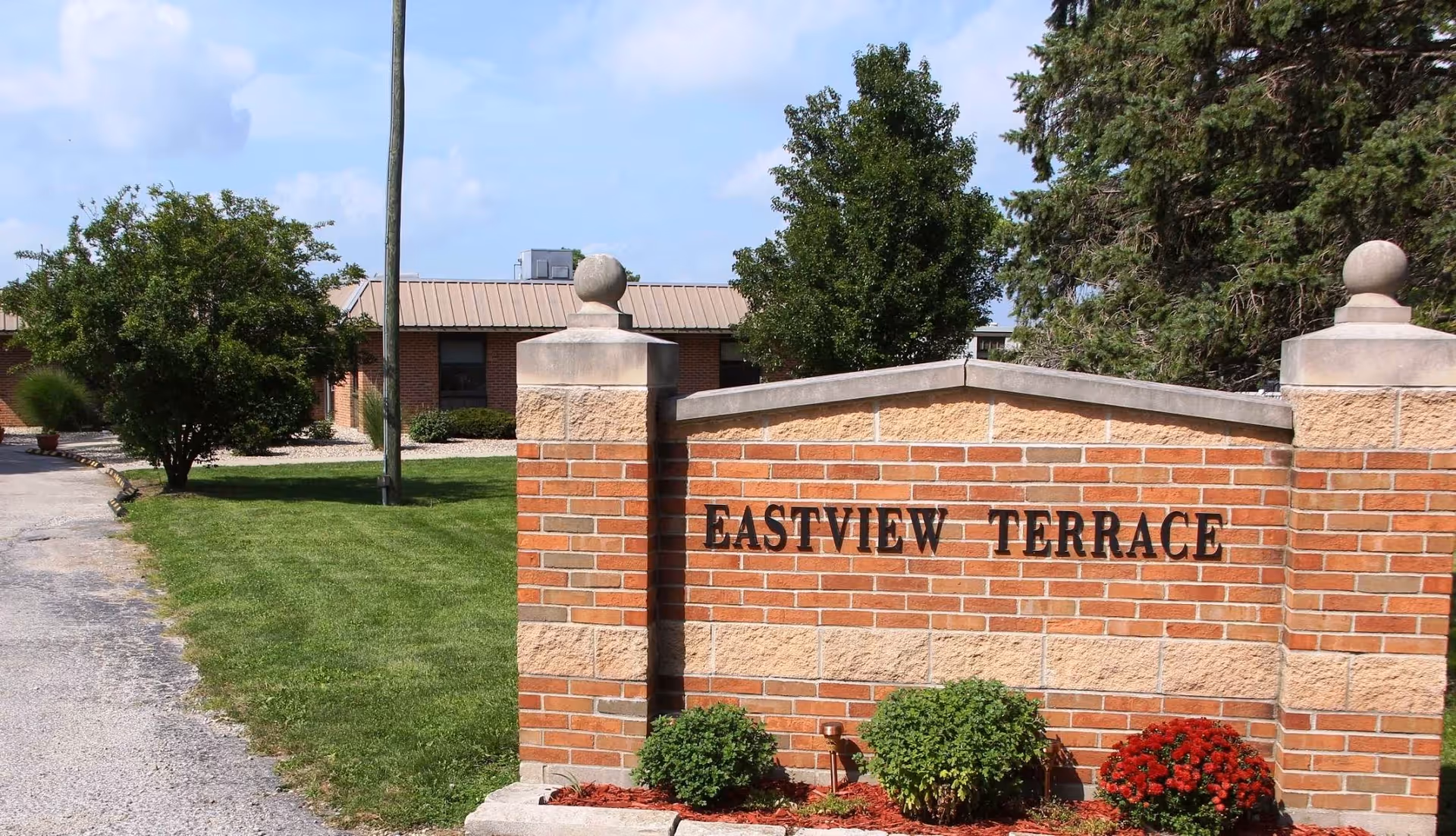 A brick sign with the text 'EASTVIEW TERRACE' in black letters, surrounded by green bushes and red flowers. Behind the sign, there is a single-story building with a tan roof and several windows, along with trees and a clear blue sky.