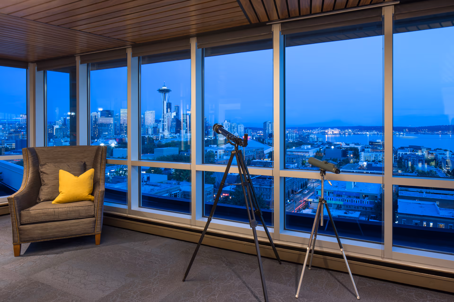 Cozy interior lounge with an armchair and two telescopes facing floor-to-ceiling windows overlooking a city skyline at dusk.