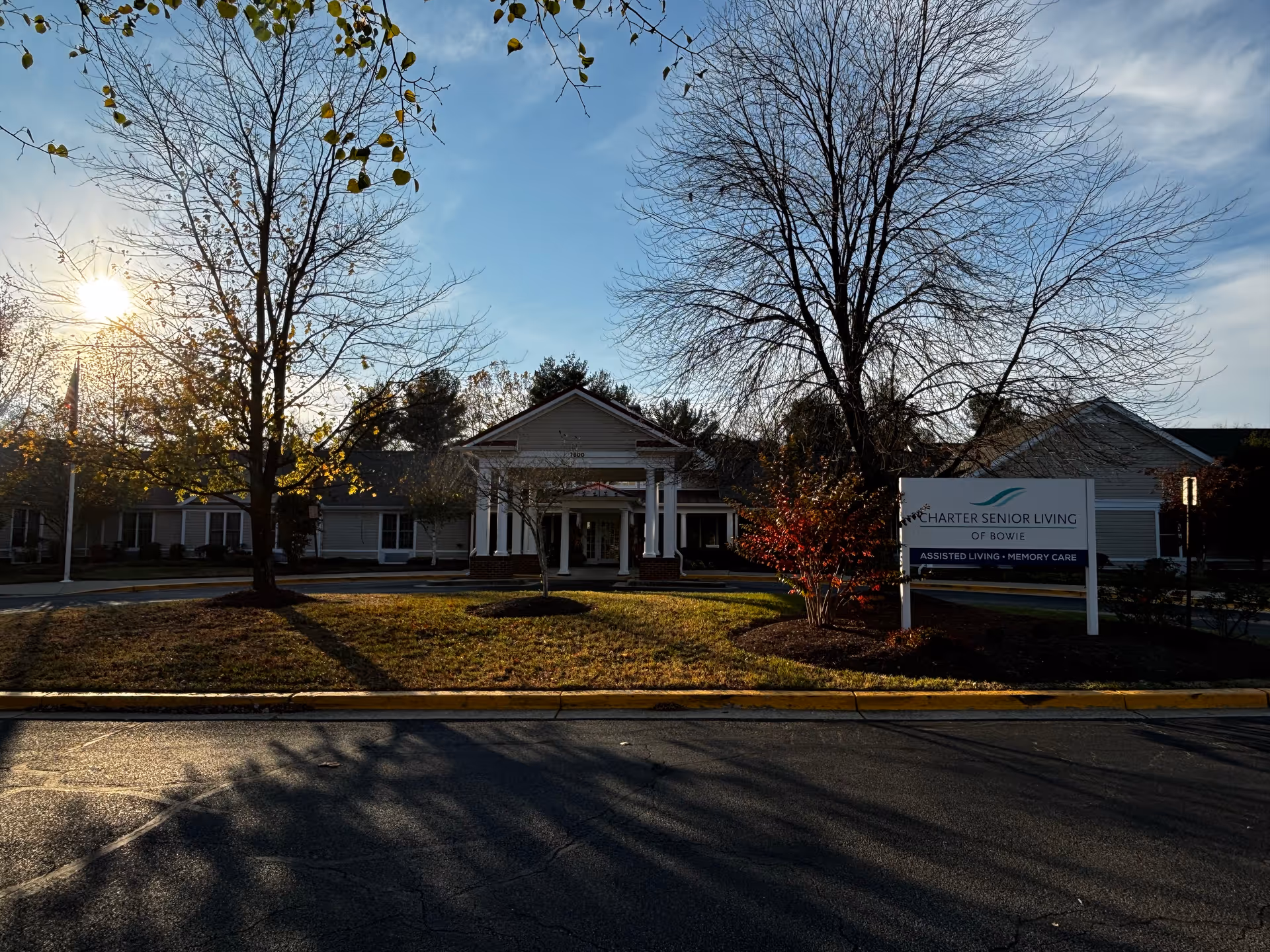 Exterior view of Charter Senior Living of Bowie building with a driveway and landscaped area featuring trees and bushes. The sun is low in the sky, casting long shadows. A sign in front reads 'Charter Senior Living of Bowie Assisted Living - Memory Care.'