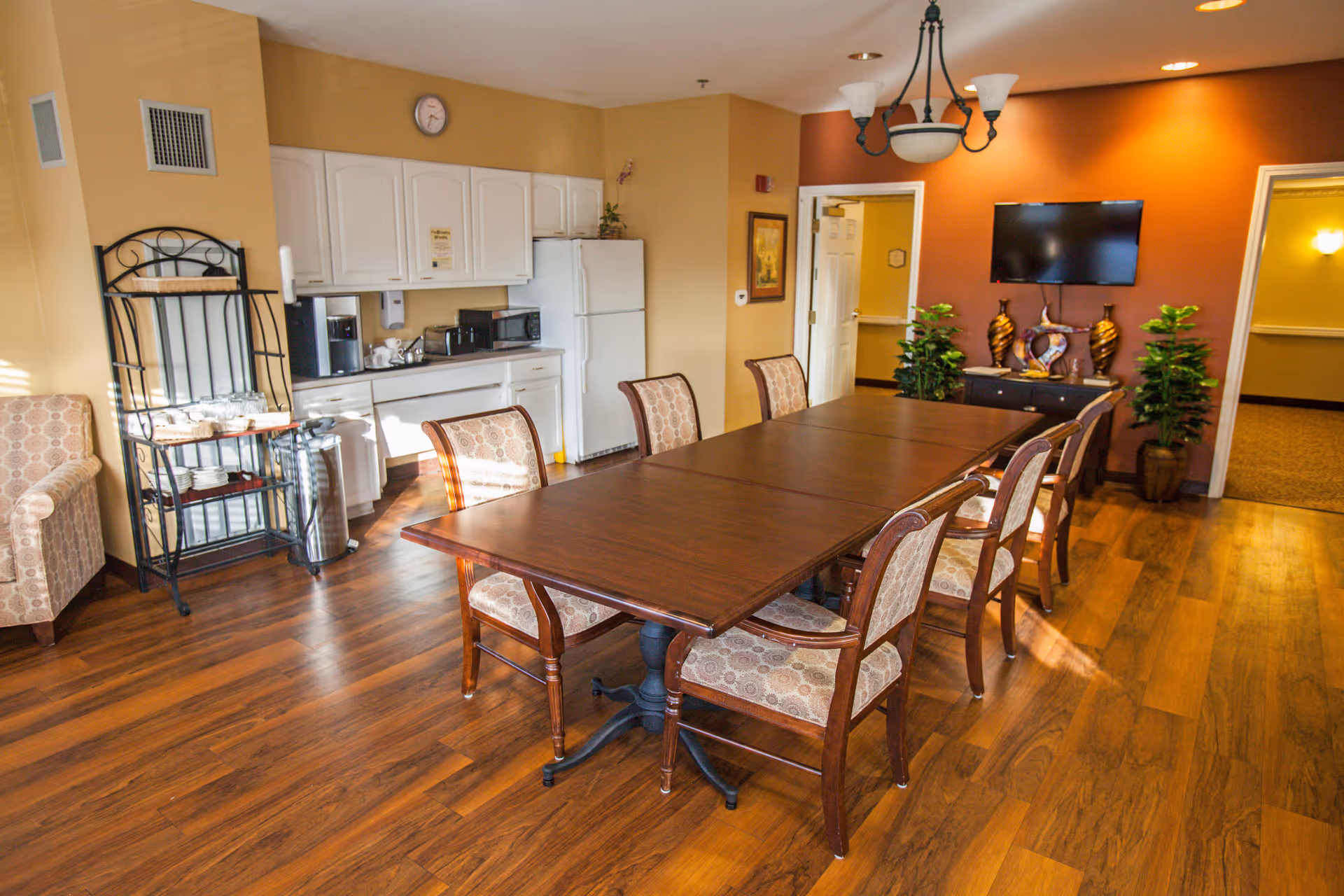 A well-lit dining room with a long wooden table surrounded by eight cushioned chairs. The room features wood flooring, a small kitchenette area with white cabinets, a refrigerator, microwave, and coffee maker. There is a decorative shelving unit with cups and saucers, a wall-mounted TV, two potted plants, and warm-colored walls with a chandelier overhead.