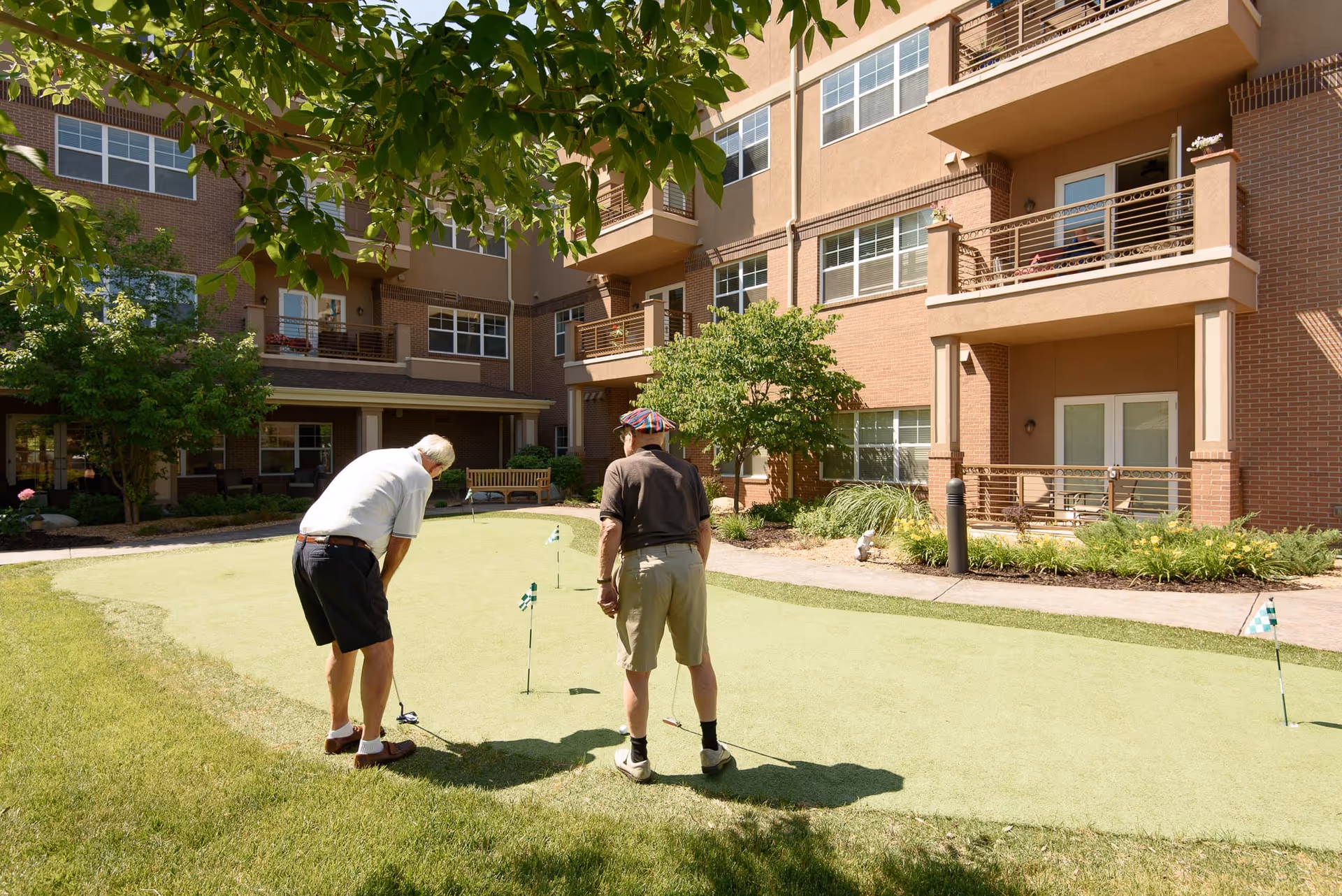 Two elderly men playing putting golf on a small green area in the courtyard of a senior living facility with a multi-story brick building in the background and trees providing shade.