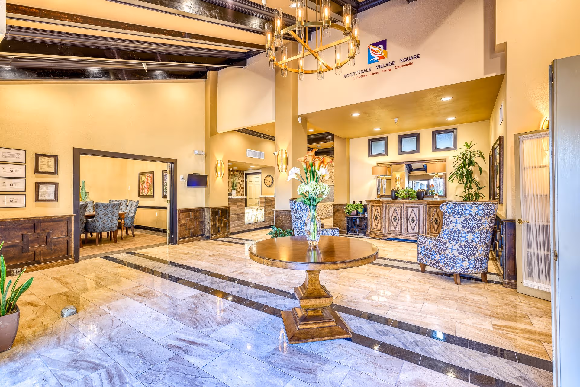 A spacious and well-lit interior lobby area of Scottsdale Village Square featuring a round wooden table with a vase of flowers in the center, patterned armchairs, a wooden sideboard with plants, and a dining area visible through an open doorway. The ceiling has exposed dark wooden beams and a modern chandelier.