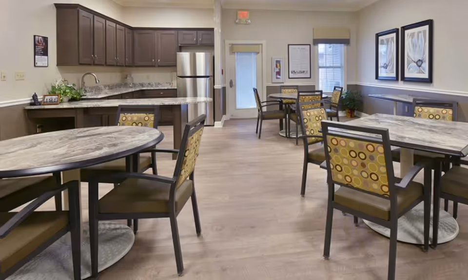 A dining area in a senior living facility featuring multiple tables with patterned chairs. In the background, there is a kitchen area with dark wooden cabinets, a granite countertop, a stainless steel refrigerator, and a sink. The room has light-colored walls, wood flooring, framed artwork, and windows with blinds.