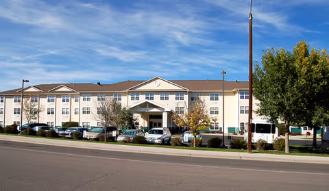 Exterior view of a three-story senior living facility building with a covered entrance, several parked cars in front, and trees on either side under a clear blue sky.