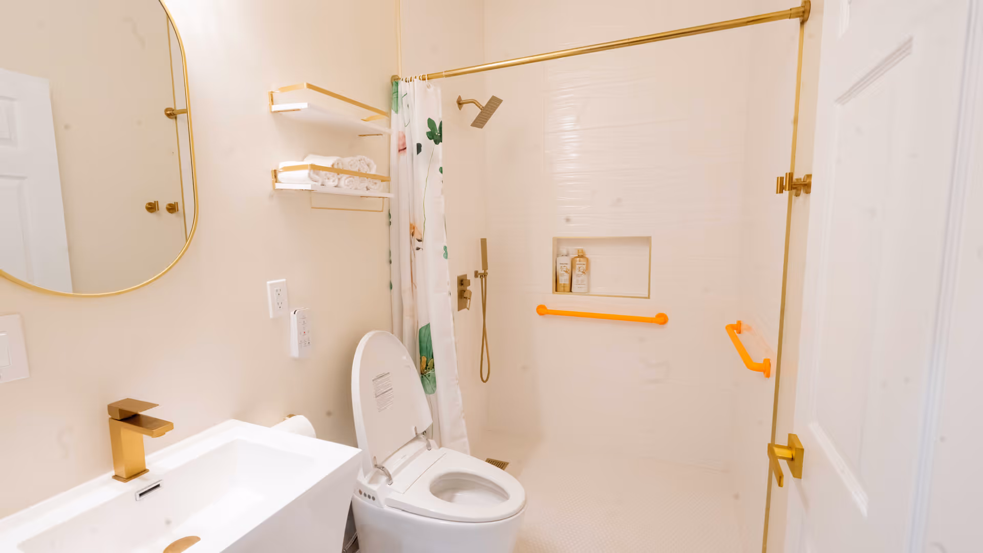 A modern bathroom with a white toilet, a white sink with a gold faucet, a large mirror with a gold frame, and a walk-in shower with a gold showerhead and orange grab bars. There are shelves with rolled white towels and a floral shower curtain.