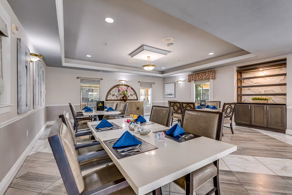 A dining room in a senior living facility with tables set for meals. The tables have white surfaces with blue folded napkins, silverware, and small flower arrangements. The room features beige walls, decorative wall art, and windows with valances. There is a ceiling light fixture and recessed lighting, along with a cabinet and decorative wall niche in the background.