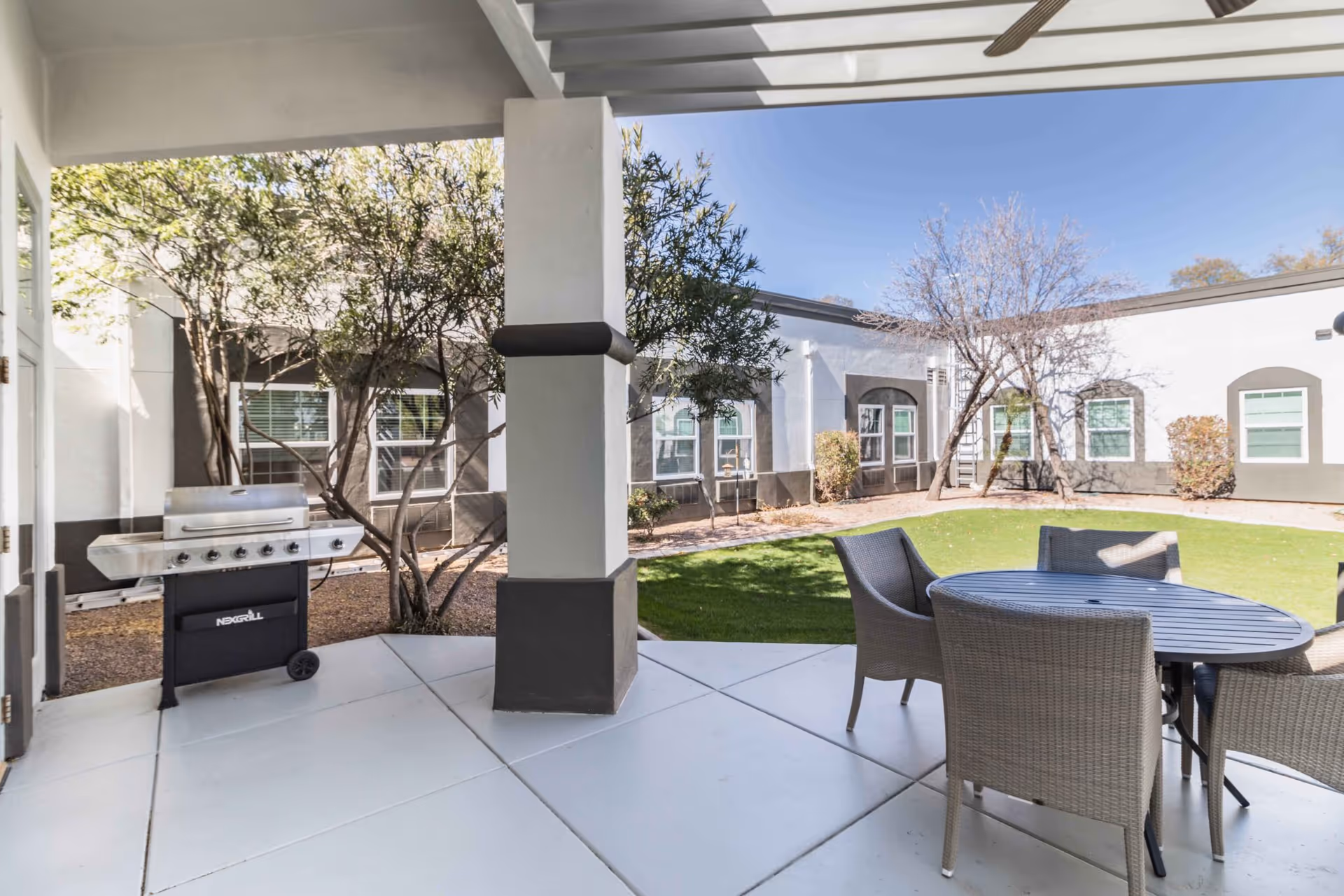 Outdoor patio area with a covered section featuring a stainless steel grill and a round table surrounded by four wicker chairs. The patio overlooks a grassy courtyard with trees and shrubs, surrounded by a white building with multiple windows.