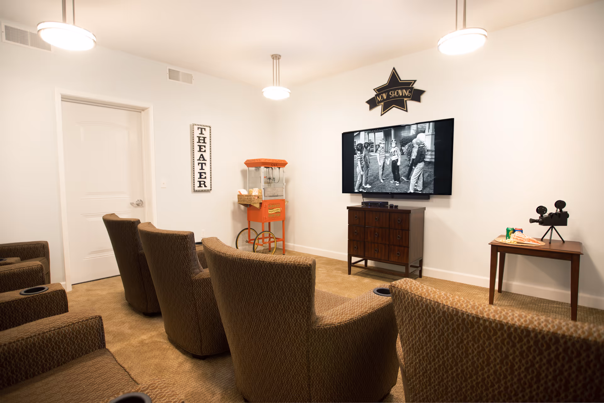 A small theater room with several upholstered recliner chairs facing a wall-mounted TV screen showing a black and white movie. The room has beige carpet, white walls, and ceiling lights. There is a vintage-style popcorn machine and a small table with a decorative film camera. A sign on the wall reads 'THEATER' and another above the TV says 'NOW SHOWING'.