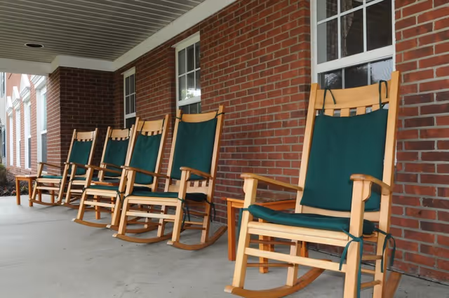 A row of wooden rocking chairs with green cushions lined up on a covered porch outside a brick building with white-trimmed windows.