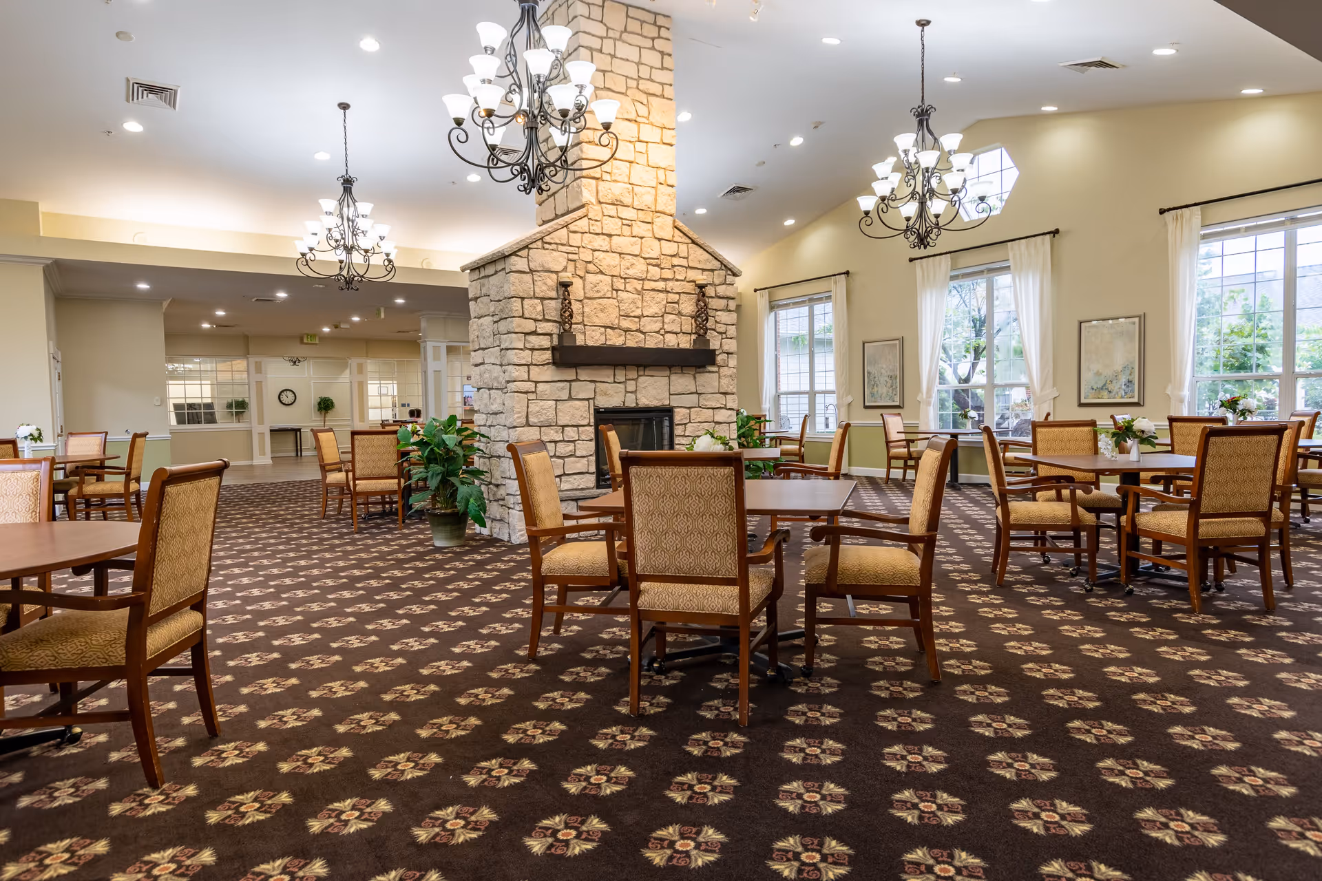 Spacious dining room with multiple wooden tables and cushioned chairs arranged around a central stone fireplace. The room features large windows with white curtains, patterned carpet, and elegant chandeliers hanging from the ceiling. Potted plants and framed artwork decorate the space, creating a warm and inviting atmosphere.