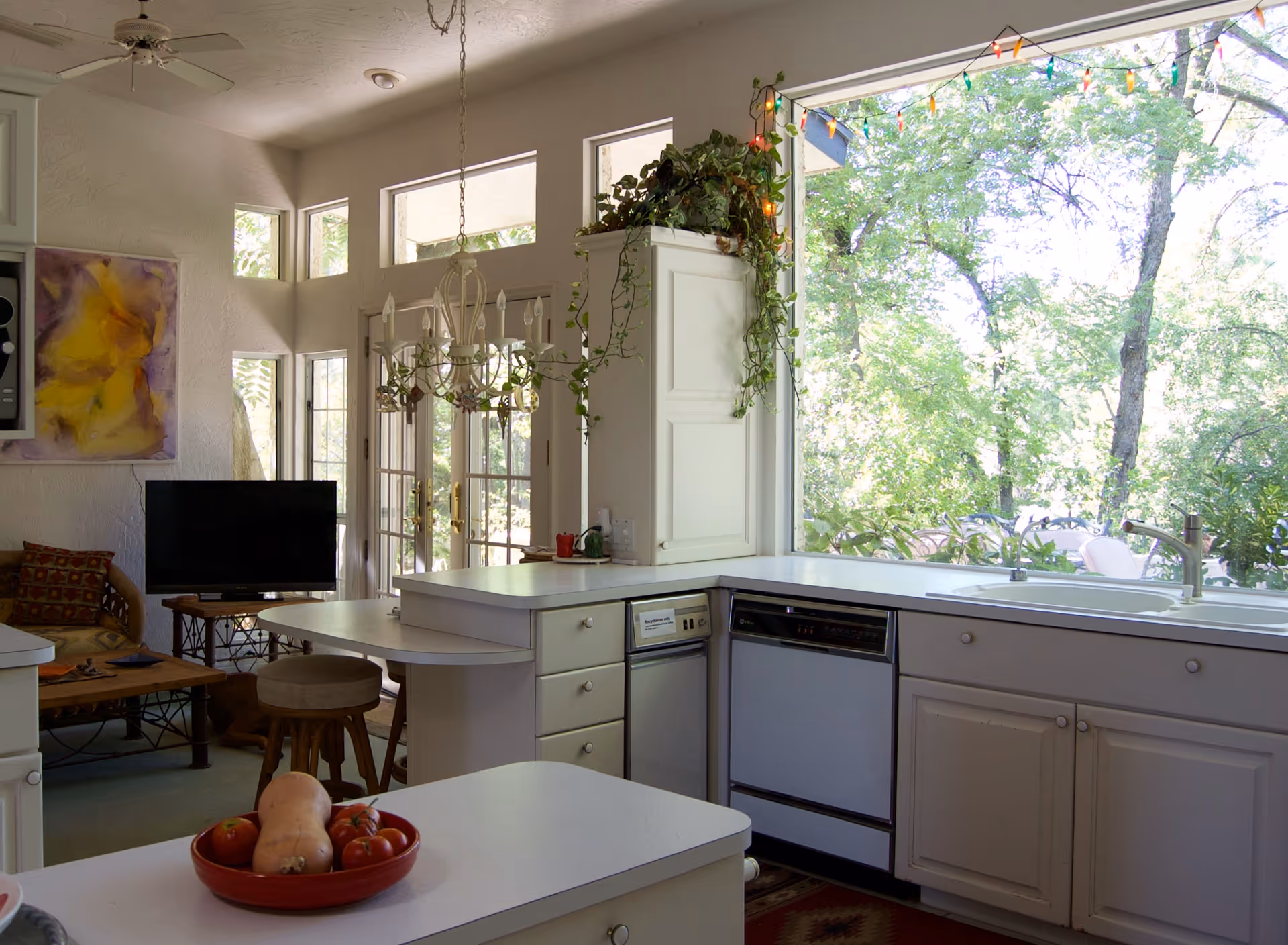 Bright kitchen area with white cabinets and countertops, a large window overlooking green trees, a sink, dishwasher, and a bowl of fresh vegetables on the counter. In the background, there is a small living area with a TV, a wooden coffee table, a stool, and a colorful abstract painting on the wall. A chandelier hangs from the ceiling near glass doors leading outside.