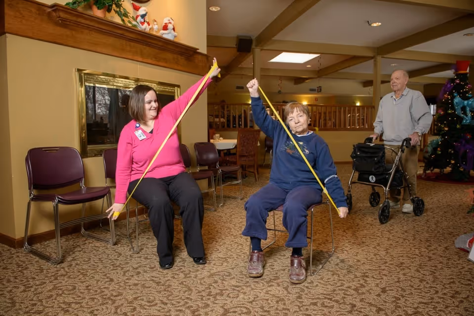 Two women seated on chairs in a common area are exercising with yellow resistance bands, while an elderly man with a walker stands nearby. The room has carpeted floors, a fireplace, and a decorated Christmas tree in the background.